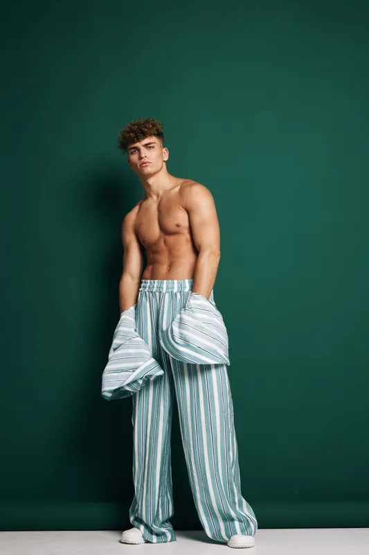 Young muscular man posing shirtless wearing striped wide pants holding a shirt standing on floor with distance to dark green wall background studio.