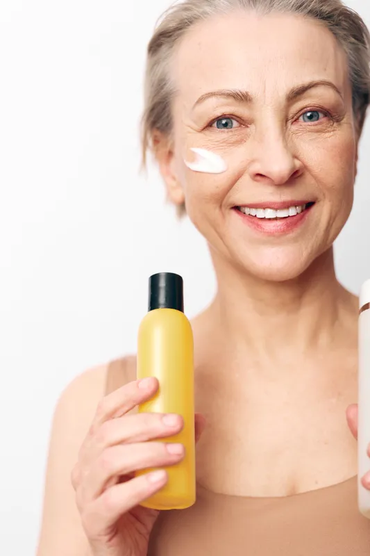 Smiling mature woman with natural skin holds yellow bottle and applies white cream to face. Close-up on white background. Concept of skincare, anti aging, wellness, and self care routine.