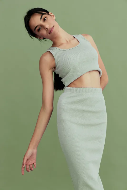 young woman wearing a sleeveless crop top and matching skirt, posing against an olive green background, contemporary fashion style with relaxed mood