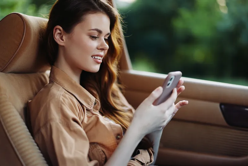 Young woman enjoying a sunny day in the car, engaging with her smartphone while seated in a stylish vehicle surrounded by a lush green background.
