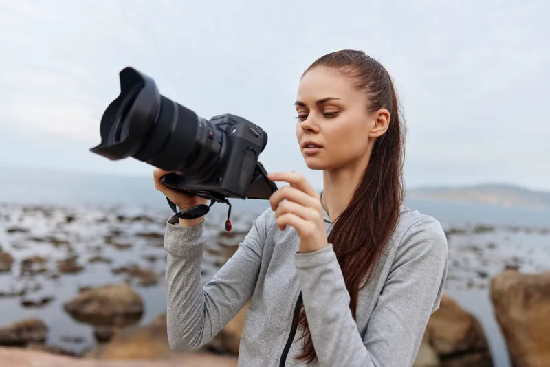 Young woman with long brown hair holding a professional camera, dressed in casual gray apparel, focused on photography by the sea at dusk