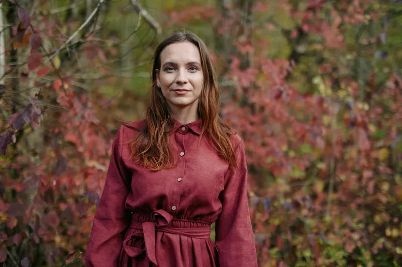 Authenticity and credibility are visible as a calm woman stands in a burgundy dress amid autumn foliage, natural light, and a serene, genuine expression in a simple, honest portrait.