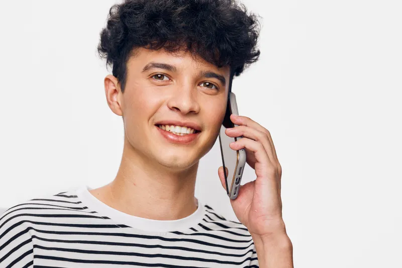 smiling man talking on smartphone with curly hair wearing striped shirt against white background, communication and technology concept