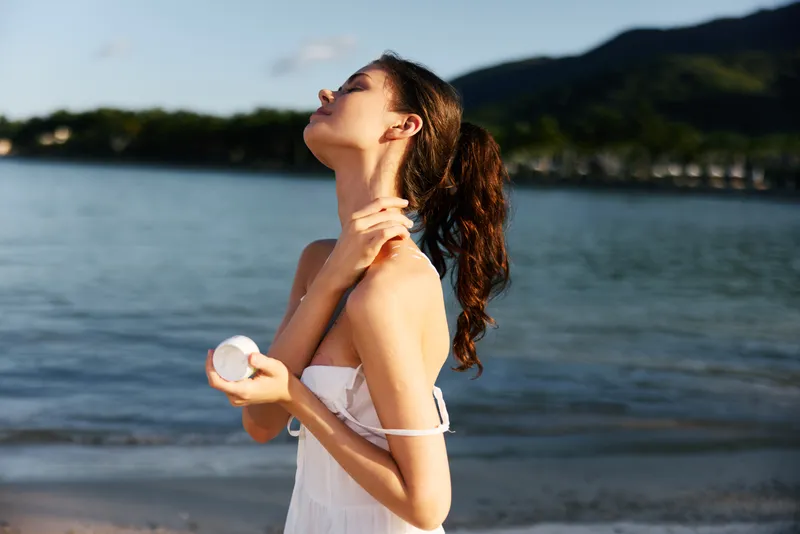 Relaxed woman applying skincare cream by the sea during a sunny summer day, enjoying natural beauty and ocean breeze.