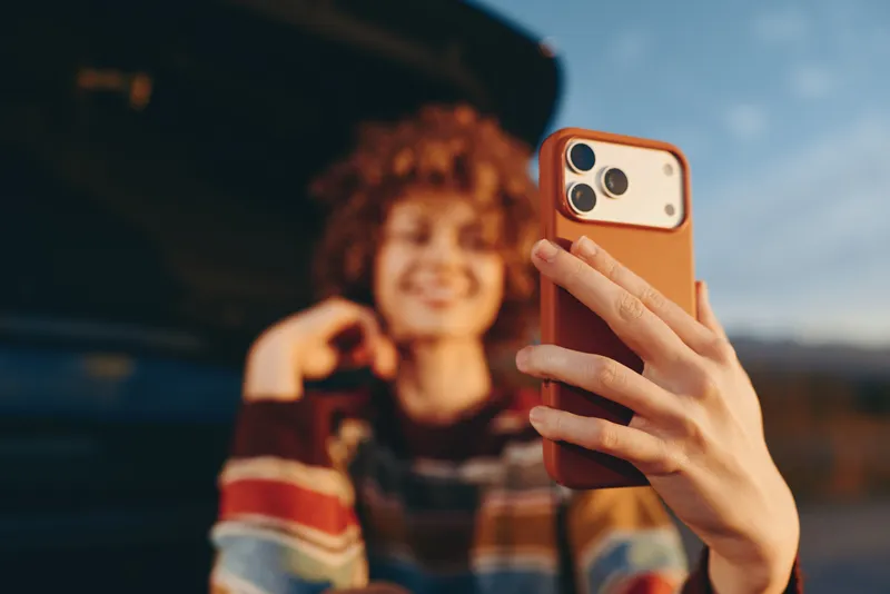 Woman does selfie with smartphone, outdoor lifestyle, hand closeup holding phone, curly hair woman smiling softly, blurred street background in sunset light, casual sweater, social media