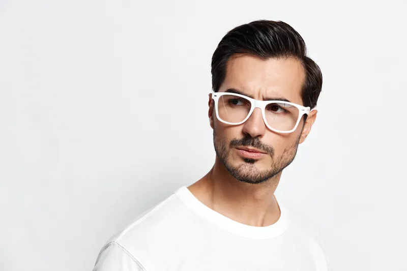 Stylish man wearing white frame glasses and a casual white t-shirt looking thoughtfully to the side against a clean, minimalistic background. Fashion concept.