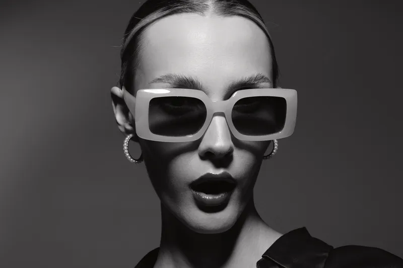 Stylish white woman with pale skin wearing large square sunglasses and hoop earrings, posing with mouth slightly open in black and white studio portrait.