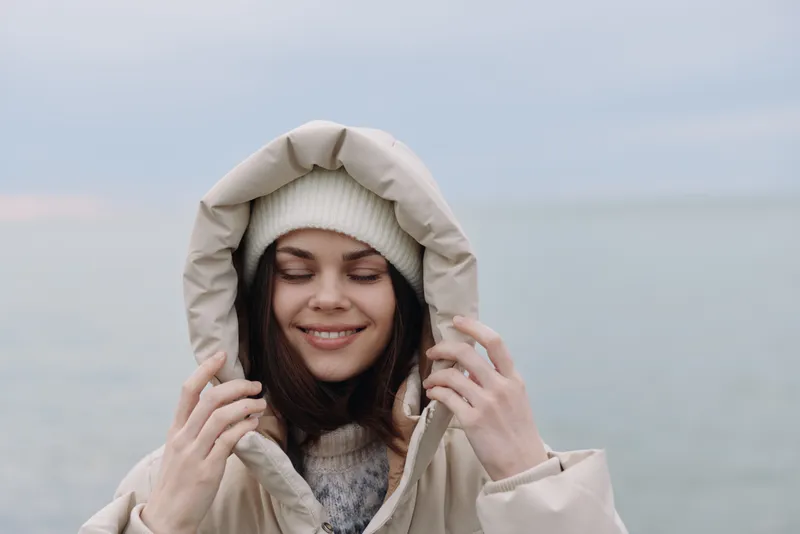 Woman wearing a warm hooded winter coat on a chilly beach, smiling softly, cozy outerwear in beige tones against a calm coastal backdrop and open horizon.