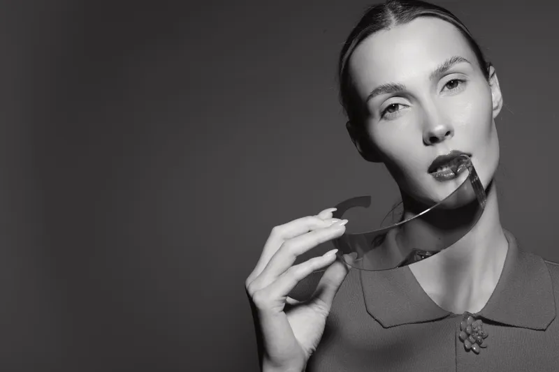 Caucasian woman with fair skin holds transparent glasses near lips, looking at camera. Black and white portrait with elegant and minimalist concept.