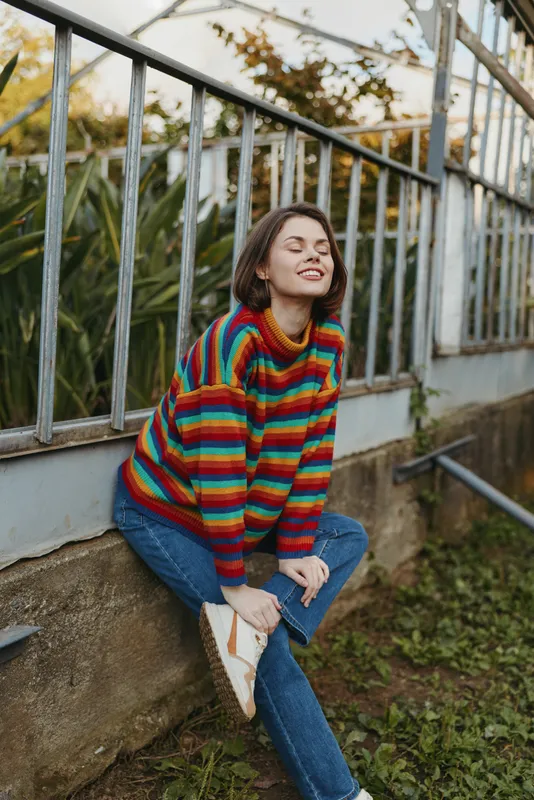Woman in colorful striped sweater and beanie sitting on low wall by metal railing outdoors, smiling with closed eyes, relaxed casual pose in urban garden or greenhouse setting.