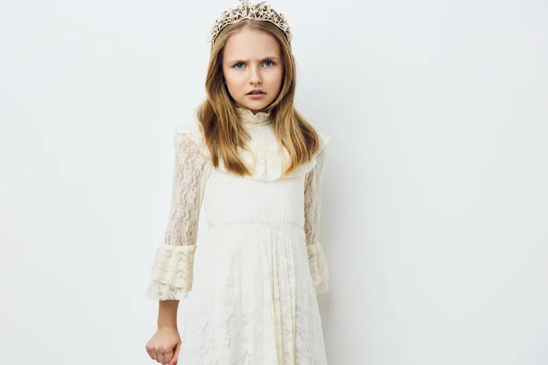 A young girl in a vintage white dress and tiara, expressing a serious emotion against a minimalistic white background Perfect for themes of childhood and royalty