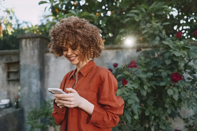 Young woman with curly hair in an orange blouse smiles while using a smartphone in a sunny garden with blooming roses and a soft sun flare
