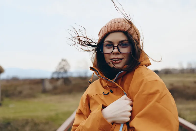 Woman outdoors lifestyle portrait, wind in air, orange coat, knit hat, glasses, smiling softly, adjusting collar, countryside field, light breeze, hair flying, relaxed posture