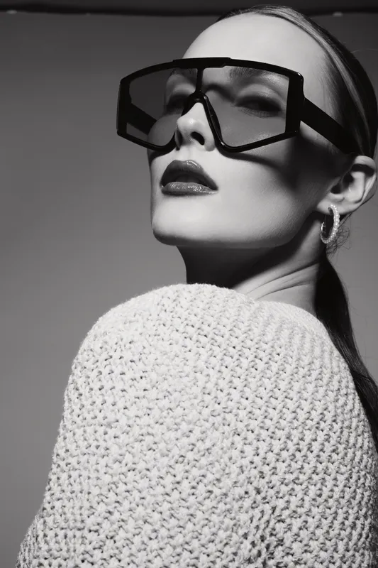 Stylish white woman with pale skin wearing oversized sunglasses and knitted sweater looks confidently in studio. Fashion portrait, modern style, elegance, and beauty concept.