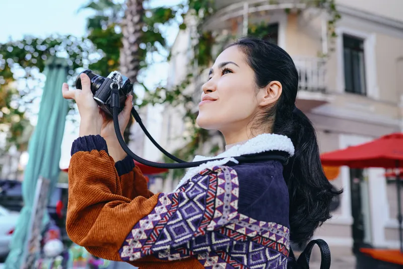Candid scene of a woman holding a DSLR camera up to shoot a lively outdoor street scene, wearing a colorful patterned jacket and scarf in bright daylight.