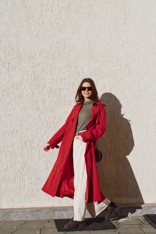 Stylish woman in a bright red coat walks beside a sunlit wall, wearing a green top and white pants, exuding confidence and casual elegance.