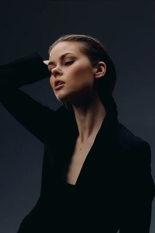 Portrait of thoughtful woman with slicked-back hair wearing black outfit against dark background, expressing contemplation and elegance.