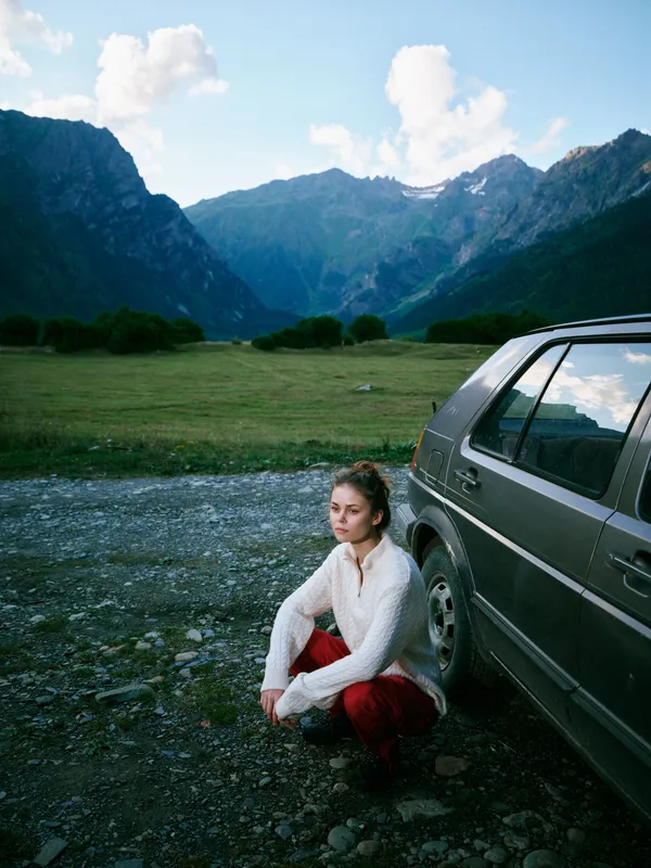 Woman crouching beside a car in a mountain valley, lush meadow, distant peaks, blue sky, tranquil outdoor travel scene, adventure and exploration in a scenic alpine landscape