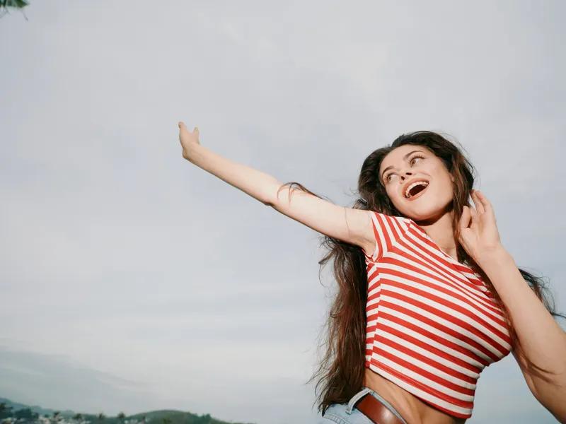 Joyful young woman in a red and white striped top raises her arms in a bright outdoor setting, smiling widely, wind in her hair, capturing carefree energy and candid charm.