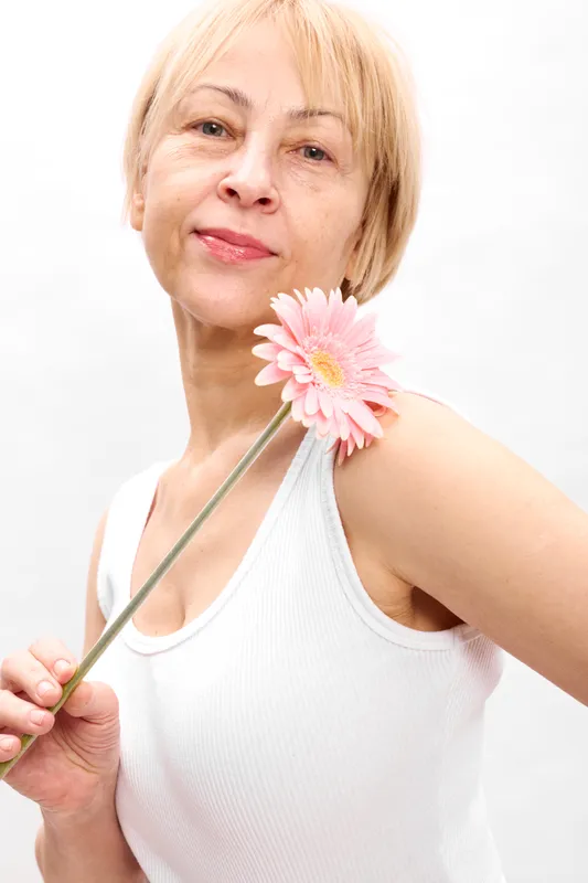 Elderly woman portrait on white background, smiling softly while holding a pink flower to her shoulder, grasping the green stem, beauty wellness relaxation concept skincare care gentle