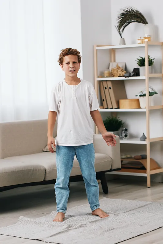 Young boy stands barefoot on a light rug in a bright living room, wearing a white t shirt and blue jeans, posing casually with relaxed posture and friendly expression.