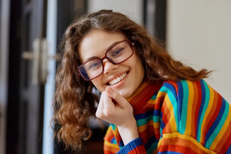 A smiling young woman with curly brown hair and red frames rests her chin on her hand while wearing a colorful striped sweater indoors.