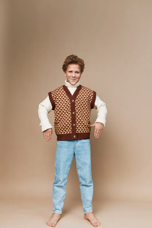 Young boy wearing a brown patterned knitted vest over a cream shirt, light blue jeans, and barefoot, posing with a confident stance against a warm tan studio backdrop.