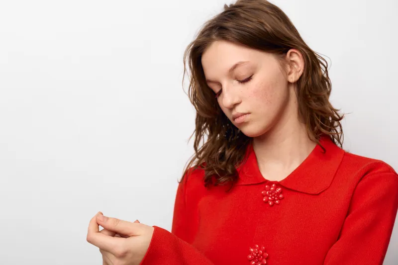 Young woman in red sweater looks down with closed eyes, holding her wrist. Studio portrait on white background. Concept of introspection, calmness, and emotional reflection.
