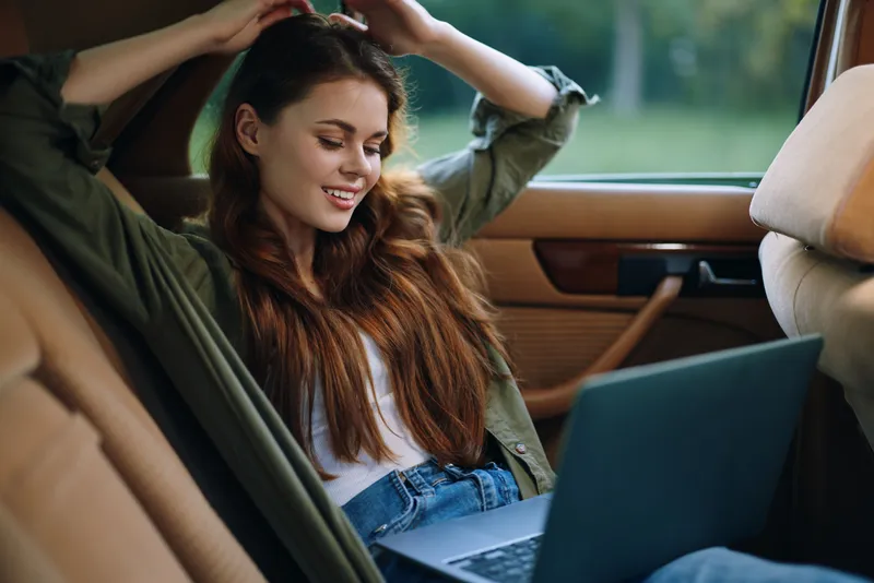 Young woman with long hair relaxing in a car, working on a laptop, wearing casual outfit, smiling happily in natural light.