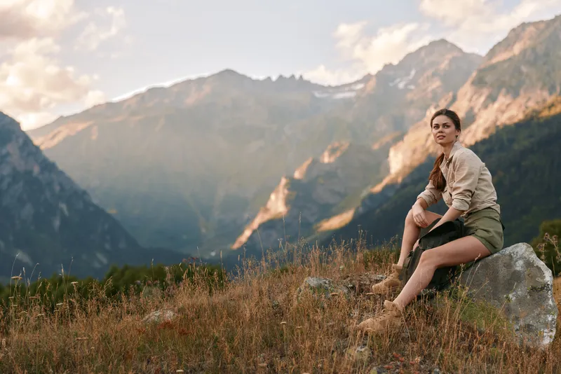 Solitary young woman sits on a rocky outcrop in a rugged mountain landscape, enjoying the fresh air and sunlit scenery with a calm contemplative expression.