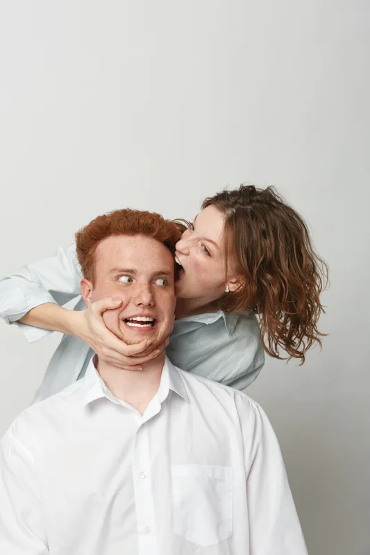 Playful young woman with curly hair pretending to bite the cheek of a surprised redhead man in white shirts. Concept of fun, friendship, humor, and lighthearted interaction between friends.
