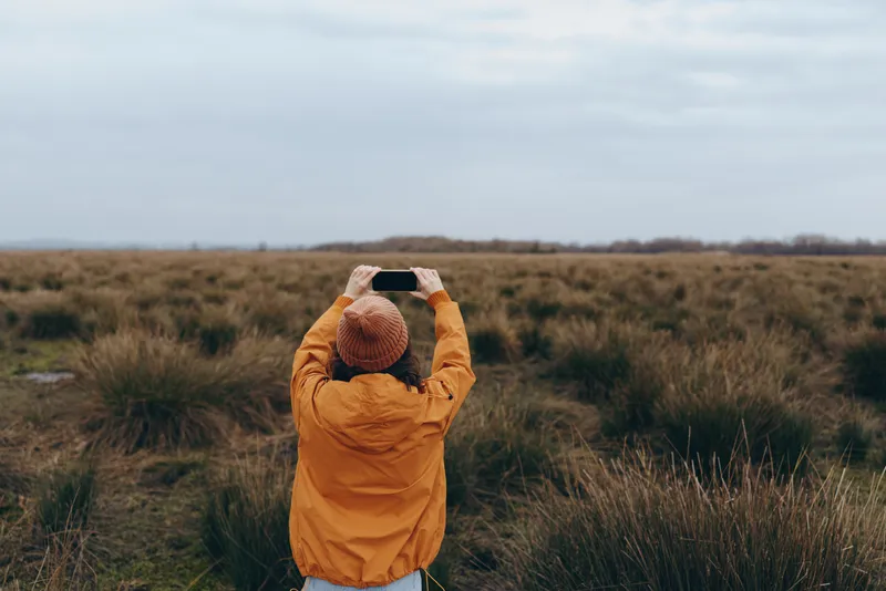Woman outdoors in a grassy field, wearing a wintercoat, holding a hat on her head, seen from behind, nature landscape under a cloudy sky, lifestyle concept, calm relaxation, soft daylight