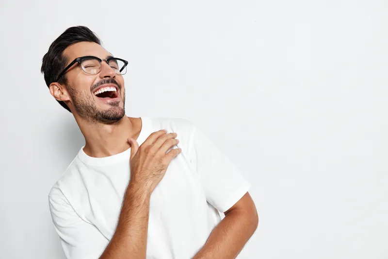 Happy joyful smiling man wearing glasses and a white t-shirt laughing while looking to the side isolated on a plain light gray background. People lifestyle concept.