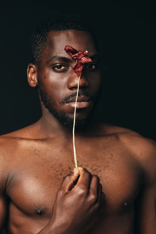 Creative conceptual portrait of African man holding dried flower in front of face with dark background. Artistic visual style with bare chest and serious expression for modern photography.
