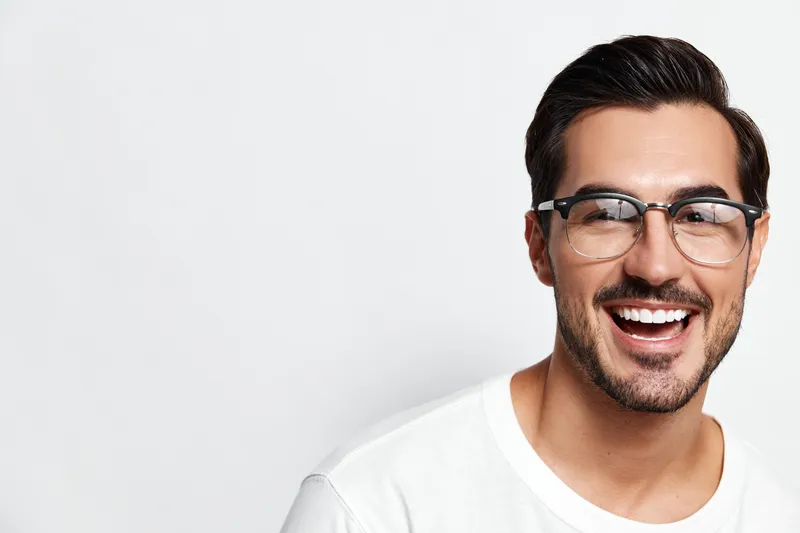 Happy smiling young caucasian man wearing glasses and a white t-shirt, looking at the camera with a cheerful expression against a light background.