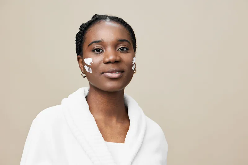 Young Black woman applying skincare product, featuring a calm expression and a cozy white robe, against a beige background, emphasizing self care and beauty