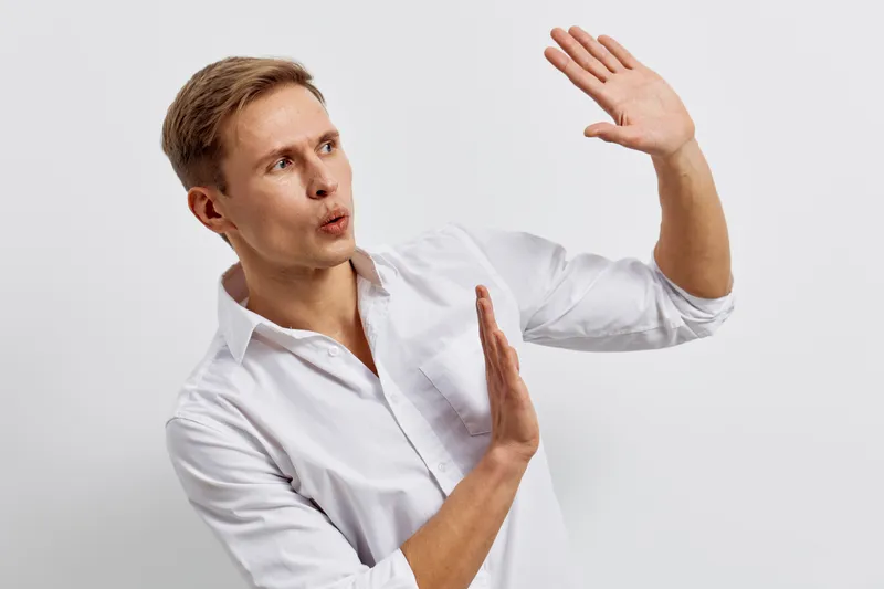 Elegant young man with blonde hair in white shirt making defensive gesture with hands, surprise or rejection expression, isolated on plain background. Studio portrait, people concept