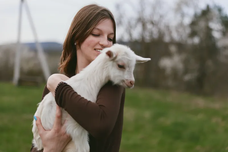Cheerful woman outdoors, cuddling a young goat kid, warm sunlight, gentle embrace, rural field backdrop, natural colors, peaceful mood, affectionate bond