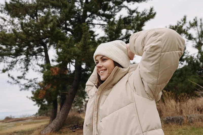 Outdoor scene with a woman in a beige puffer jacket and knit beanie adjusting her hood in a winter landscape, with trees and an open field behind for a calm seasonal portrait.