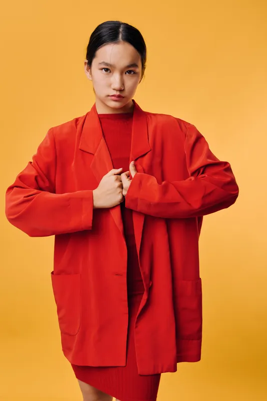 Young woman in red oversized jacket and dress posing on yellow background with serious expression. Fashion style with minimal makeup and natural hair for modern portrait.
