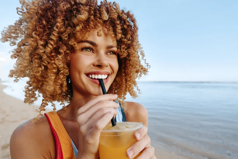 Smiling woman with curly hair on a sunny beach holds a jar drink with straw and ice, relaxing by the ocean during summer vacation, carefree seaside lifestyle.