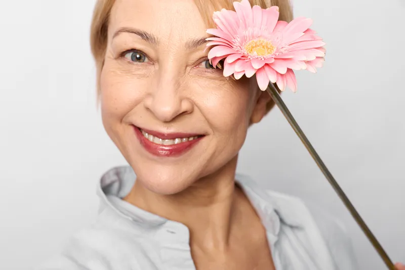 Woman elderly smiling portrait holding pink gerbera flower closeup on light background with spring mood soft focus friendly expression for beauty and wellness