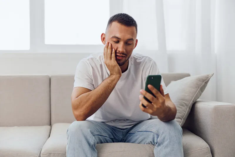 Thoughtful man using smartphone while relaxing on a cozy sofa, portraying a moment of reflection and everyday life. Ideal for lifestyle and technology themes.
