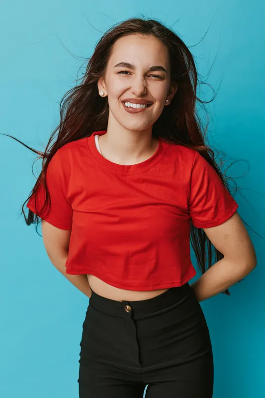 Young woman smiling with closed eyes wearing red crop top and black pants standing against blue background. Long hair flowing and casual joyful expression for lifestyle portrait.