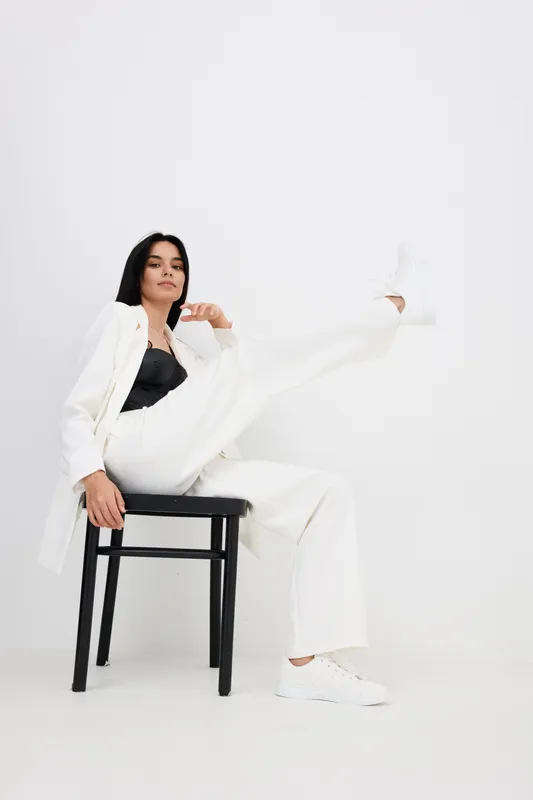 Stylish woman in white suit and black top sitting on black chair with one leg raised, wearing white sneakers, modern minimalistic studio background and confident pose.