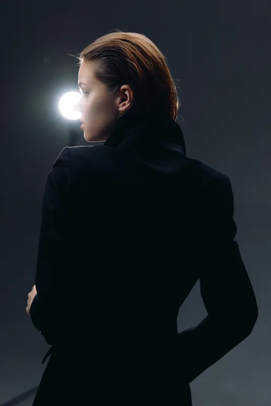 Woman in black coat standing with back turned in dramatic low light portrait with dark background.