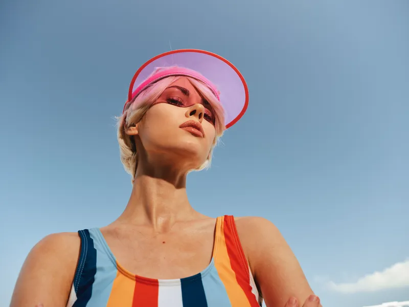 Confident model posing outdoors in bright sunlight wearing a colorful striped swimsuit and pink visor against a clear blue sky.