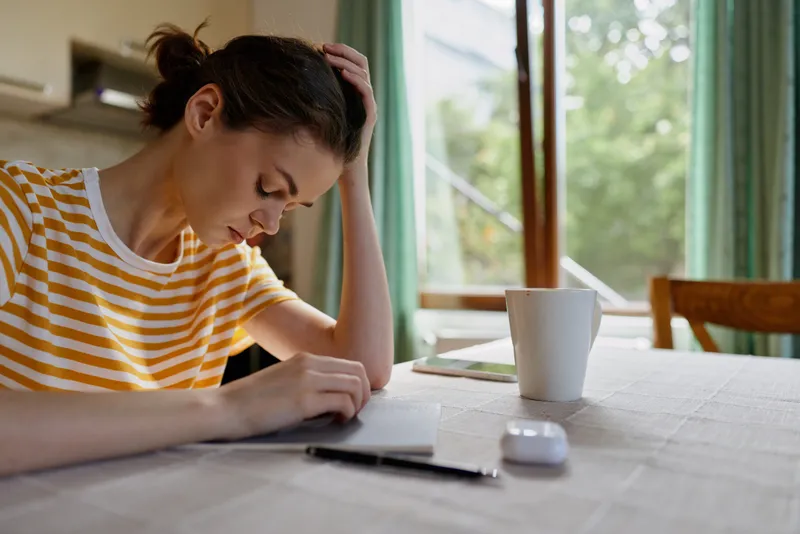 A woman sits at a sunlit table in a home, writing in a notebook with a mug nearby, conveying calm focus, daily life, and quiet work at home.