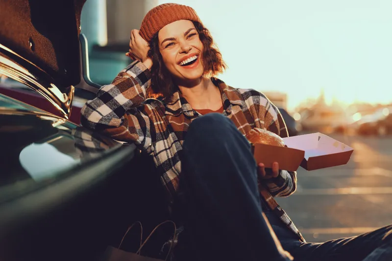 Woman by car holding pizza, laughter and beanie at sunset in urban parking, candid moment with golden hour glow, authenticity, mindful living and emotional storytelling.