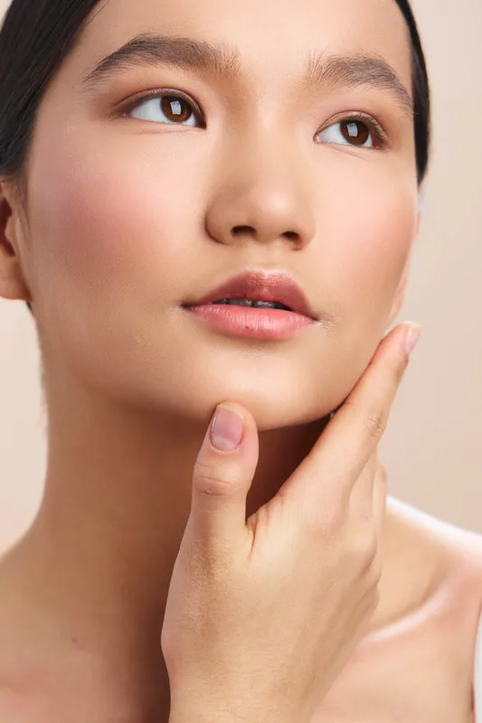 Asian woman appears in a close up, calm and thoughtful, gently touching her smooth skin, highlighting a skincare and beauty focus with soft, flattering lighting.